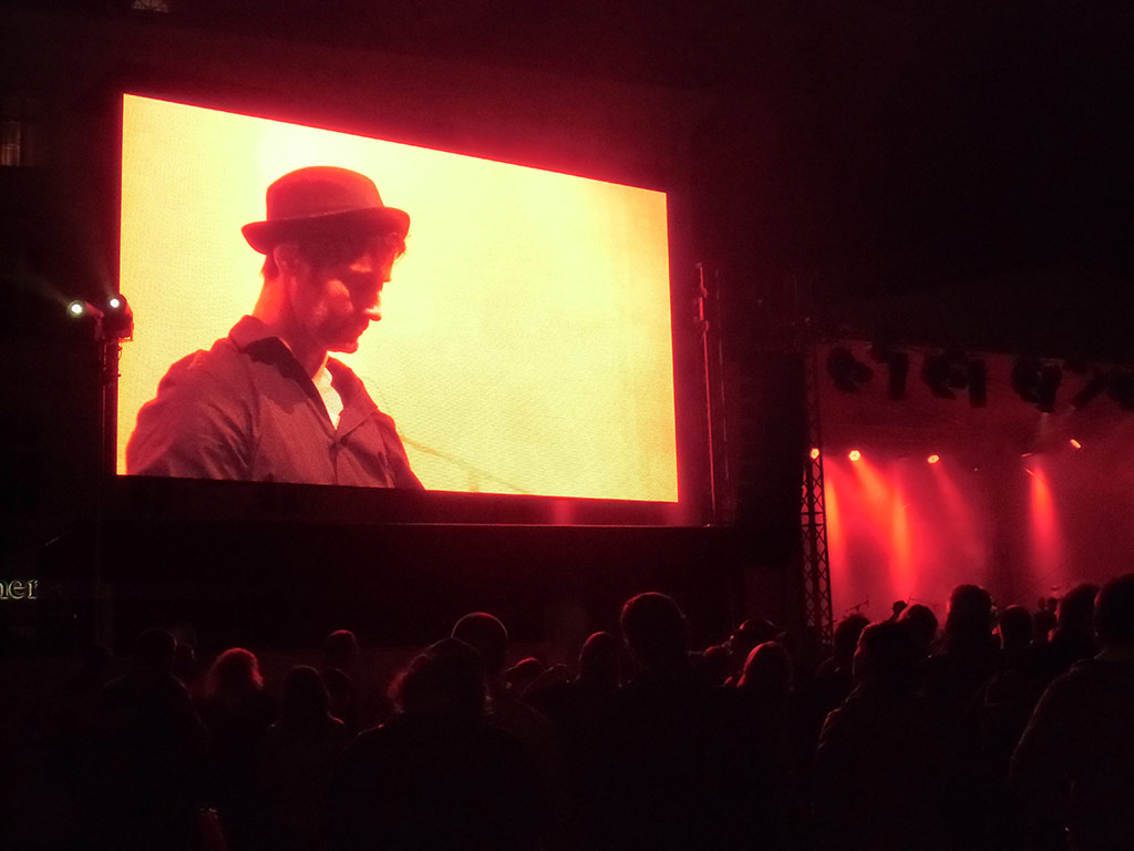 Large outdoor screen at Lichtfest 2014 displaying a close-up of a musician in a hat performing on stage under red-orange lighting, with an audience gathered below at night during an outdoor concert.