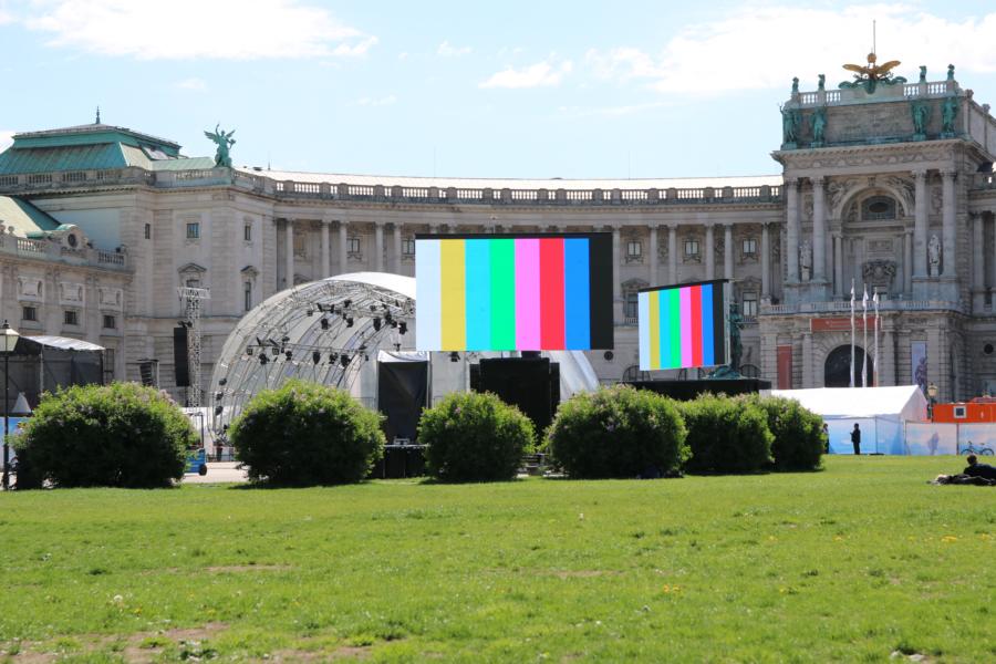 Digital screens displaying colorful test bars are set up on a green lawn in front of a historic building with arched windows, stone statues, and trimmed bushes under a clear blue sky. The modern screens contrast with the classical architecture, creating a striking visual scene.