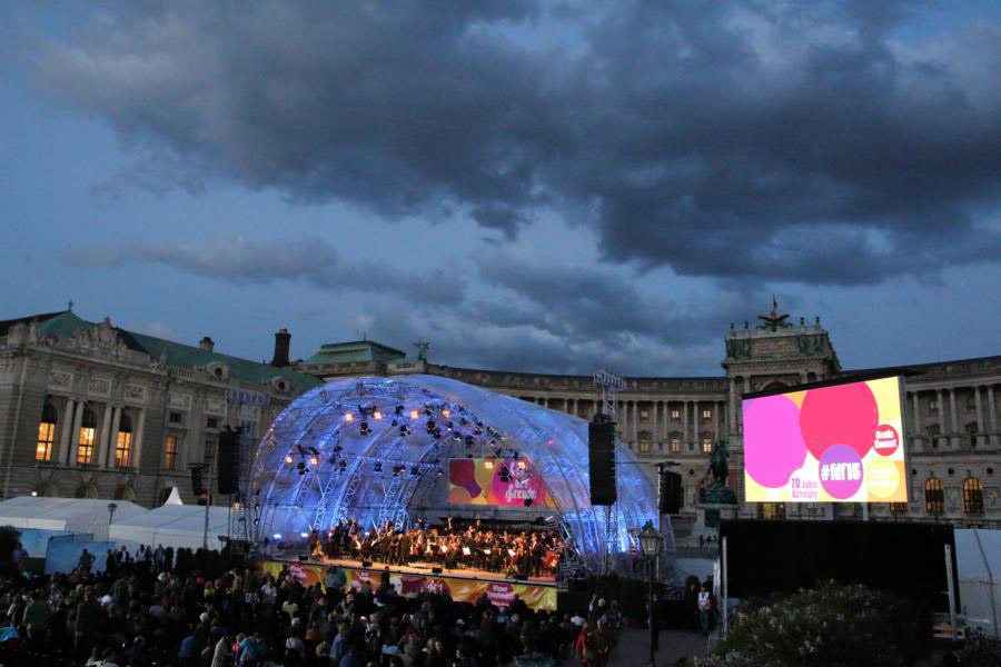 Outdoor concert at dusk in front of historic building with crowd watching illuminated stage and large screen displaying colorful graphics against dramatic cloudy sky.