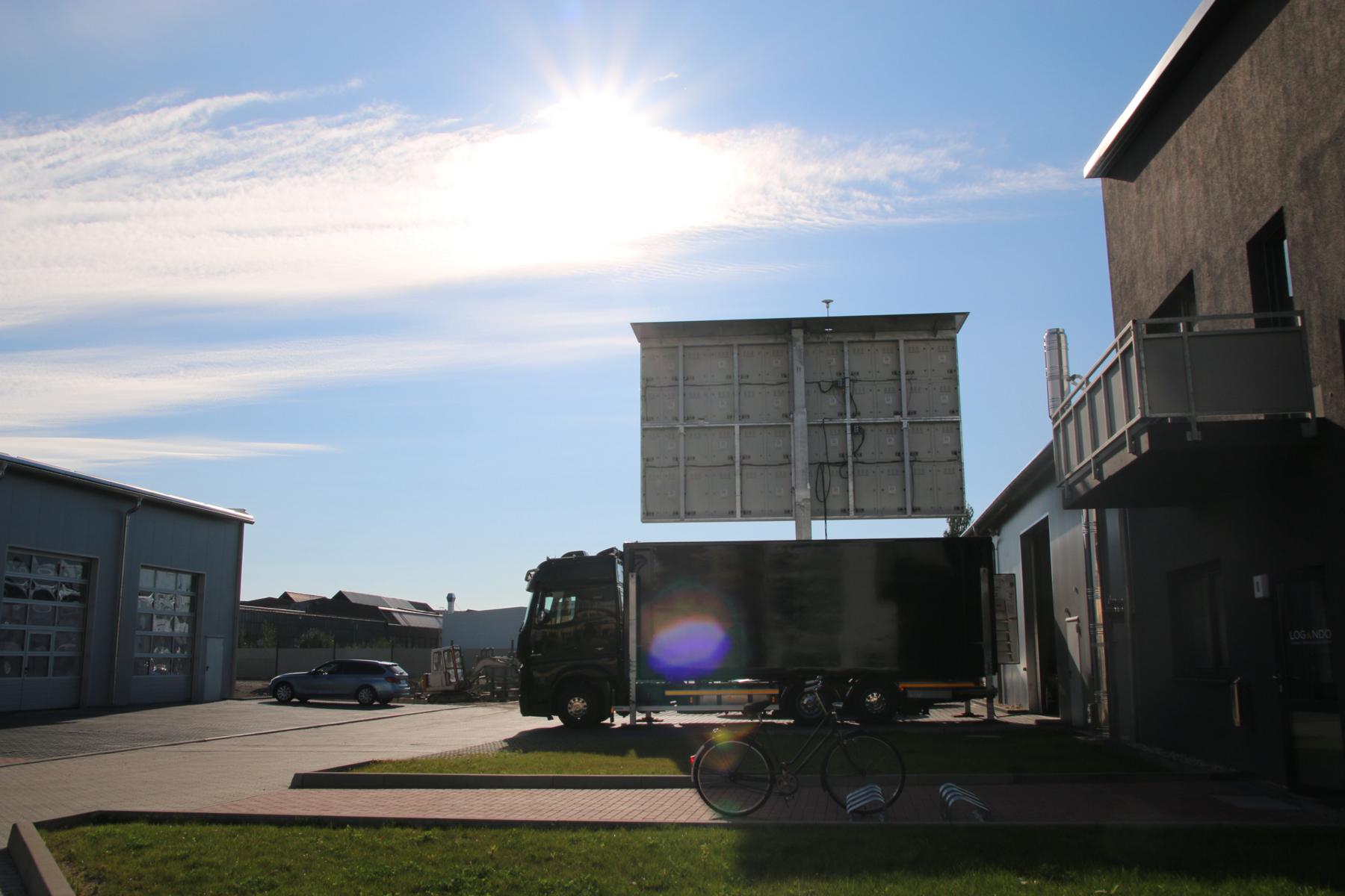 Black pickup truck parked outside industrial buildings on a sunny day, with a bicycle on the grass in the foreground and a large billboard behind the truck. Bright sunlight casts clear shadows.