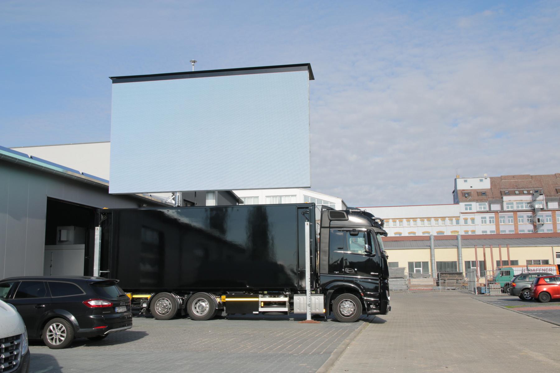 Large black mobile advertising truck with a blank raised rectangular LED screen parked in an industrial area beside cars and warehouses under a partly cloudy sky.