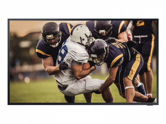 Three football players tackle an opponent on a grassy field, highlighting intense sports action—ideal for displaying on the Samsung The Terrace 75 Zoll Outdoor 4K QLED-TV, available for rent.