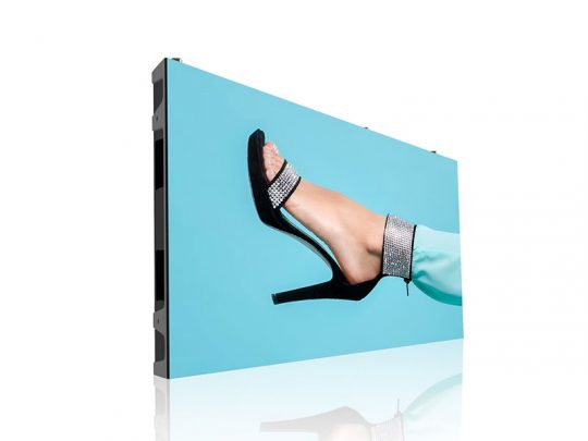 Large LED display screen featuring a close-up of a foot wearing a black rhinestone high-heeled sandal and teal pants against a vibrant blue background. Fashion footwear product image optimized for style and elegance.