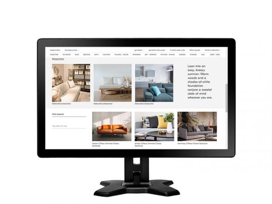 Computer monitor displaying a modern furniture website with images of contemporary sofas, sectionals, and neutral-tone living room setups. Navigation menus are visible at the top, and a short product description appears on the right side of the screen.