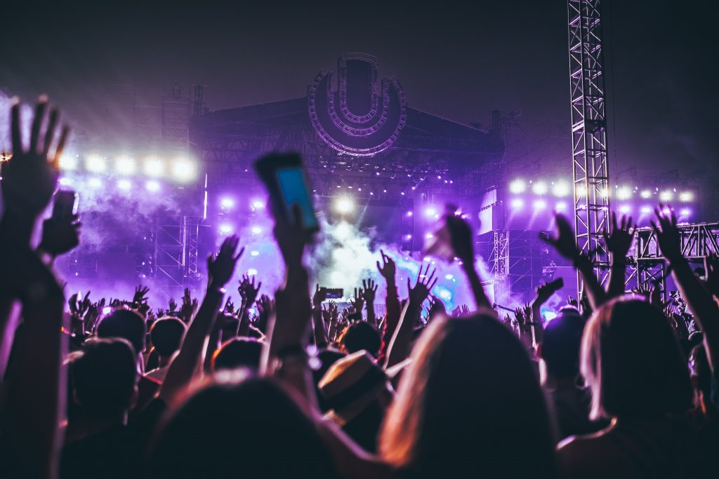 Mehrere Menschen mit erhobenen Händen feiern vor einer hell erleuchteten Konzertbühne bei Nacht. Die Bühne ist in lila Licht und Nebeleffekte getaucht. VIP-Pakete für dieses Musikfestival können gemietet oder gekauft werden, um ein unvergessliches Live-Erlebnis zu genießen.