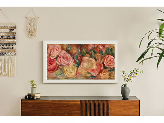 43 Zoll Samsung The Frame QLED-TV in 4K/UHD an beiger Wand montiert, wirkt wie ein Bilderrahmen; darunter Holzsideboard mit Pflanze, Büchern und Vase - idealer Designer-Fernseher als stilvolle Dekoration zum Kaufen oder Mieten für Ihr Zuhause.
