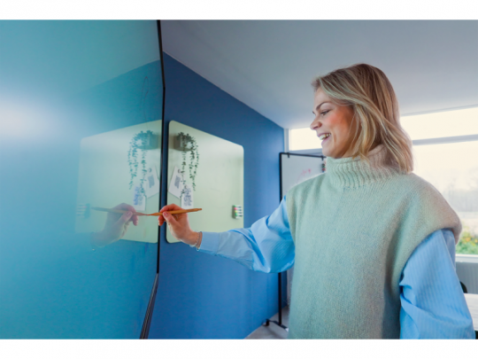 Eine Frau lächelt und bedient ein 55 Zoll Touchdisplay - Legamaster EVOLVE 2 ETX-5540 (Neuware) - in einem modernen, hell erleuchteten Raum mit blauen Wänden; ideales interaktives Whiteboard für Besprechungen oder Präsentationen, perfekt zum Kaufen.