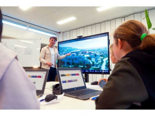 Eine Lehrkraft nutzt das 65 Zoll Touchdisplay Legamaster EXPLORE 2 EXP-6510 (Neuware) in einem modernen Klassenzimmer, um digitale Inhalte zu präsentieren. Mehrere Schüler sitzen mit Laptops vor dem interaktiven Display. Das Produkt ist zum Kaufen oder Mieten erhältlich.