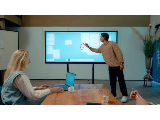 Im Besprechungsraum verwendet eine Person einen Stylus am 92 Zoll Touchdisplay Legamaster Horizon HRZ-9200 (Neuware), um "kaufen"-Optionen zu markieren; daneben macht eine weitere Person Notizen am Laptop. Das große interaktive Whiteboard eignet sich ideal für Präsentationen und Zusammenarbeit im Büro.