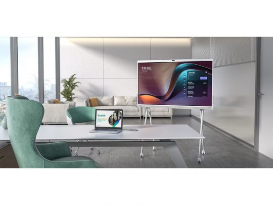 65 Zoll All-in-One Videokonferenzgerät Yealink MeetingBoard (Neuware) auf Standfuß in modernem Büro; im Vordergrund Laptop mit Website zum Kaufen oder Mieten, grüne Stühle, großes Fenster und Sofa im Hintergrund.